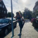 Lisette die tijdens het hardlopen een rek oefening doet bij een lantaarnpaal.