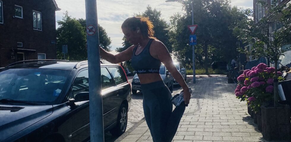 Lisette die tijdens het hardlopen een rek oefening doet bij een lantaarnpaal.
